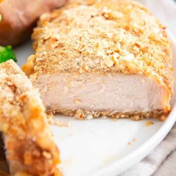 A piece of breaded and baked chicken breast is served on a white plate with broccoli and a sweet potato in the background. A fork is placed beside the plate on a striped cloth.