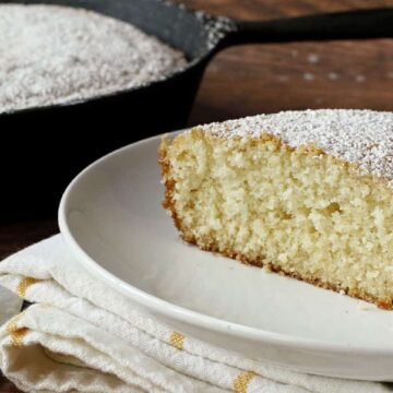 A slice of cake dusted with powdered sugar sits on a white plate, placed on a folded cloth napkin. Behind it, the rest of the cake is in a black skillet. A metal serving utensil is partially visible on the left.