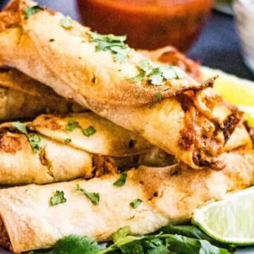 A plate of crispy taquitos stacked on top of each other, garnished with chopped cilantro. Lime wedges are on the side. A bowl of red sauce and a bowl of white sauce are partially visible in the background.