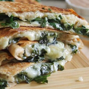 A stack of spinach and cheese quesadillas is placed on a wooden board. The quesadillas are cut into triangular pieces, with melted cheese and spinach visible. A small bowl of dipping sauce is in the background.