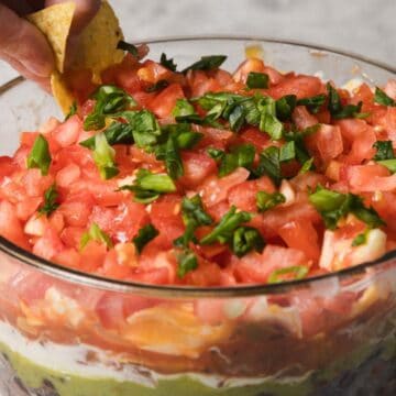 A clear glass bowl is filled with layered dip, including visible layers of beans, guacamole, sour cream, cheese, tomatoes, and chopped green onions on top. A hand is dipping a tortilla chip into the dip. A bowl of chips is nearby.