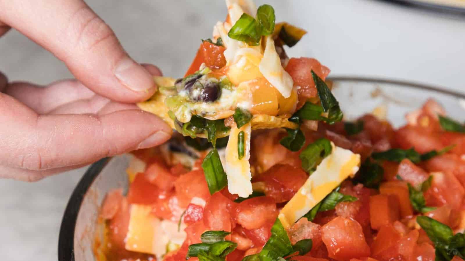 A close-up of a hand holding a tortilla chip with layers of cheese, tomatoes, black beans, and chopped green onions. The chip is angled above a bowl filled with the same mixture of ingredients. The background is slightly blurred.