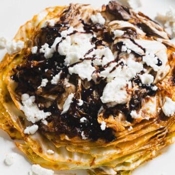 A stack of pancakes topped with crumbled cheese and drizzled with chocolate syrup sits on a white plate. The pancakes appear to have a golden-brown texture, and some cheese pieces are scattered around.