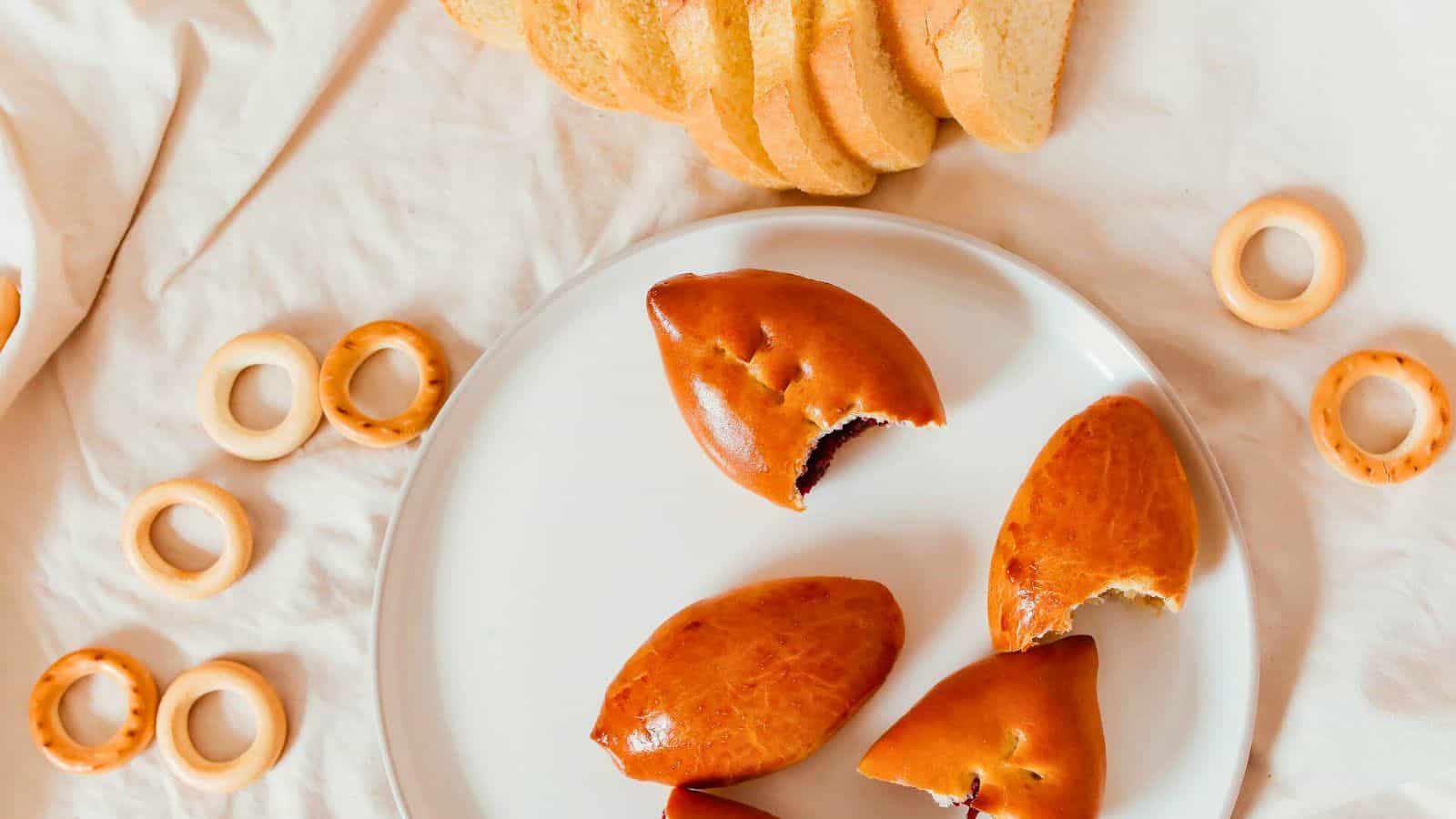 A white plate with four baked pastries, some of which have been bitten, sits on a cloth surface. Surrounding the plate are several small, round bread rings, and slices of bread are visible at the top of the image.