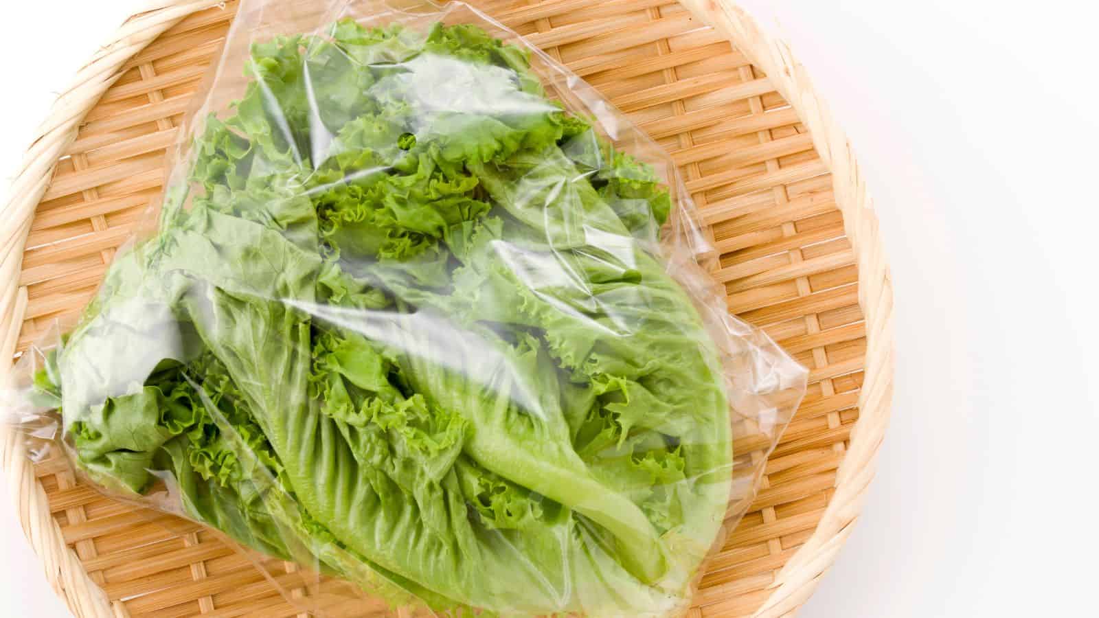 A plastic bag containing fresh green lettuce is placed on a round, woven bamboo tray. The lettuce leaves are visible through the clear packaging, with a white background surrounding the tray.