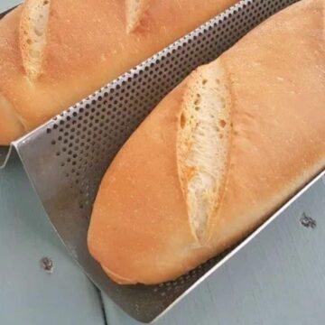 Two freshly baked homemade baguettes are placed on a perforated baking tray. The easy breads have a golden crust with three diagonal slashes on top. The tray is set on a light blue wooden surface.