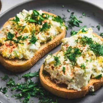 Two slices of bread topped with egg salad and garnished with fresh herbs are placed on a gray plate. A small dish with lemon slices is partially visible in the background on the left. The surface below is light-colored.