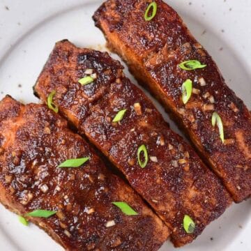 Three seasoned grilled salmon fillets, perfect for any Gordon Ramsay fan, are neatly arranged on a white scalloped plate. Garnished with chopped scallions for a pop of color, this dish is sure to impress. The plate is elegantly set on a light-colored fabric surface.