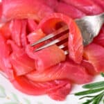 Close-up of a plate with thinly sliced smoked salmon, delicately pink and slightly curled. A fork is partially inserted into the luscious beetroot cured salmon. A sprig of rosemary graces the edge of the plate, adding an aromatic touch.
