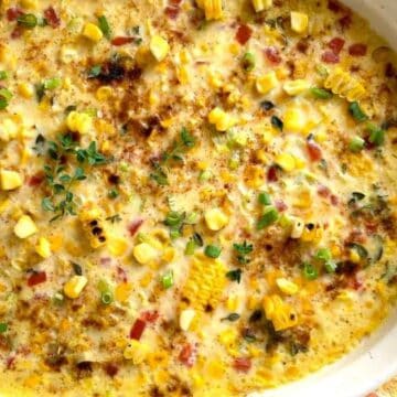 A baked casserole dish filled with a creamy mixture containing corn kernels, diced red peppers, and herbs. The top is seasoned and lightly browned, garnished with small green leaves. The dish is set on a wooden surface.