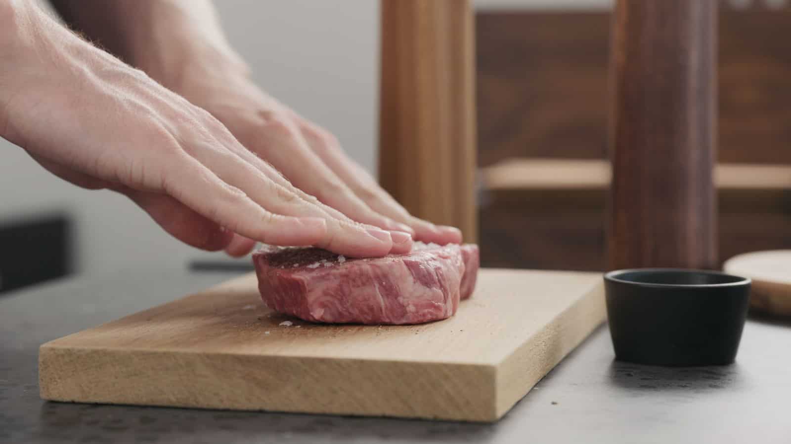 A person presses down on a raw steak with both hands on a wooden cutting board. A small black bowl is placed nearby on the gray countertop. The background is blurred, showcasing a wooden object.