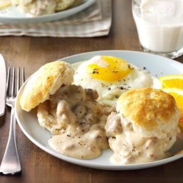 A plate of biscuits topped with gravy is served alongside a sunny-side-up egg and two orange slices. A knife and fork are placed beside the plate on a wooden table. A glass of milk is in the background.