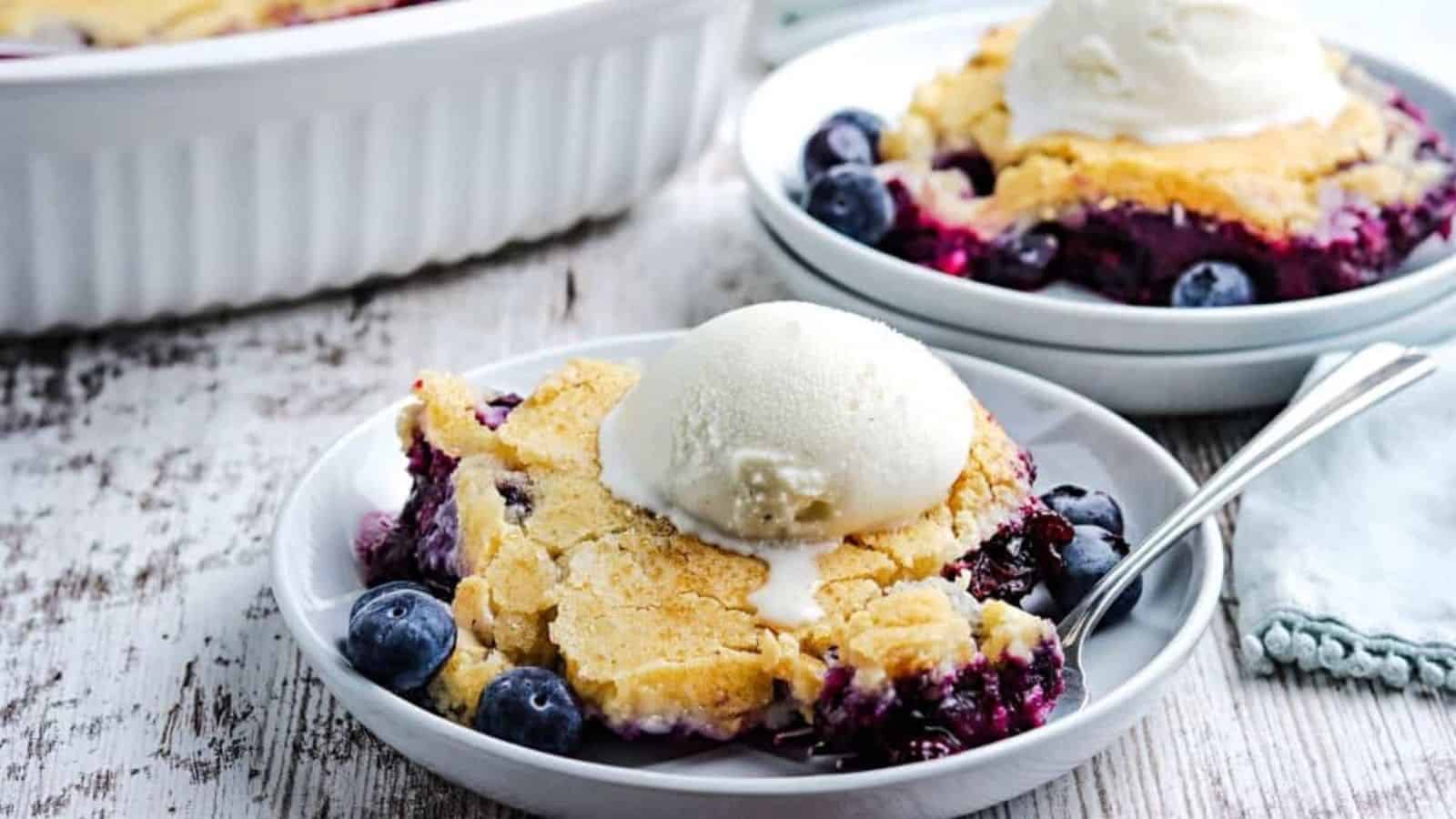 A plate with blueberry cobbler, a nostalgic treat topped with a scoop of vanilla ice cream and fresh blueberries. A spoon rests on the plate. Another similar dish and a baking dish are in the background, all placed on a wooden surface.