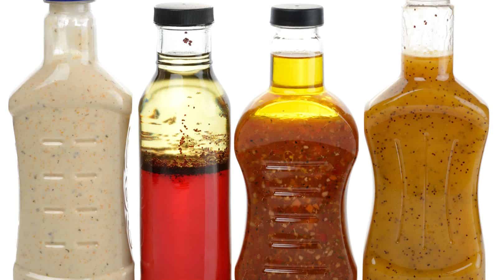 Four bottles of salad dressing are lined up against a white background. The dressings appear to be a creamy dressing, a red vinaigrette, a dressing with visible herbs and spices, and a mustard-colored vinaigrette.