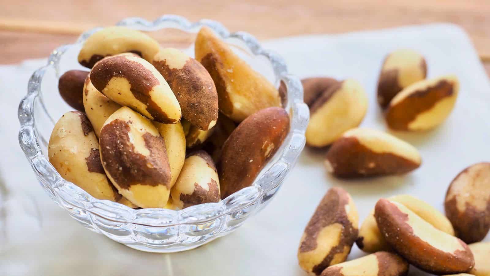 A glass bowl filled with Brazil nuts sits on a light-colored surface. Some Brazil nuts are scattered outside the bowl. The nuts are roughly oval-shaped with brown and cream-colored shells. The background is softly blurred.
