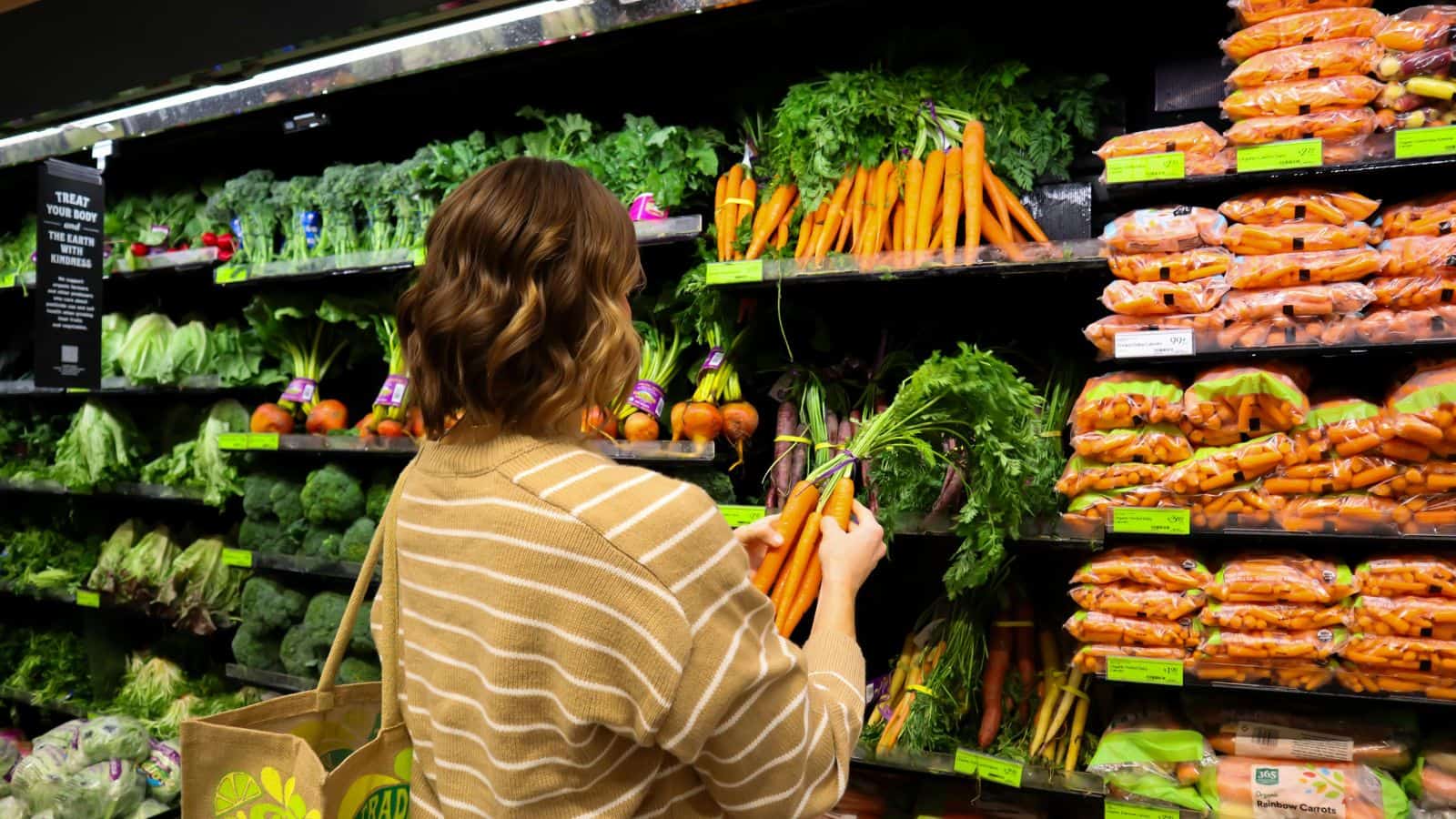 A person wearing a striped sweater selects fresh carrots from a grocery store produce section. They hold a reusable shopping bag. Surrounding shelves are stocked with various vegetables, including packaged carrots and leafy greens.