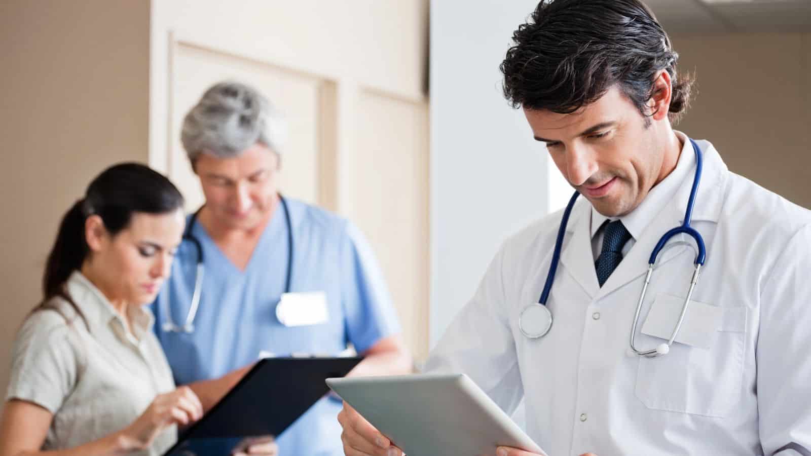 A doctor in a white coat with a stethoscope reviews a tablet in the foreground. Behind, a woman and a man in scrubs are looking at clipboards. The setting appears to be a medical facility or hospital.