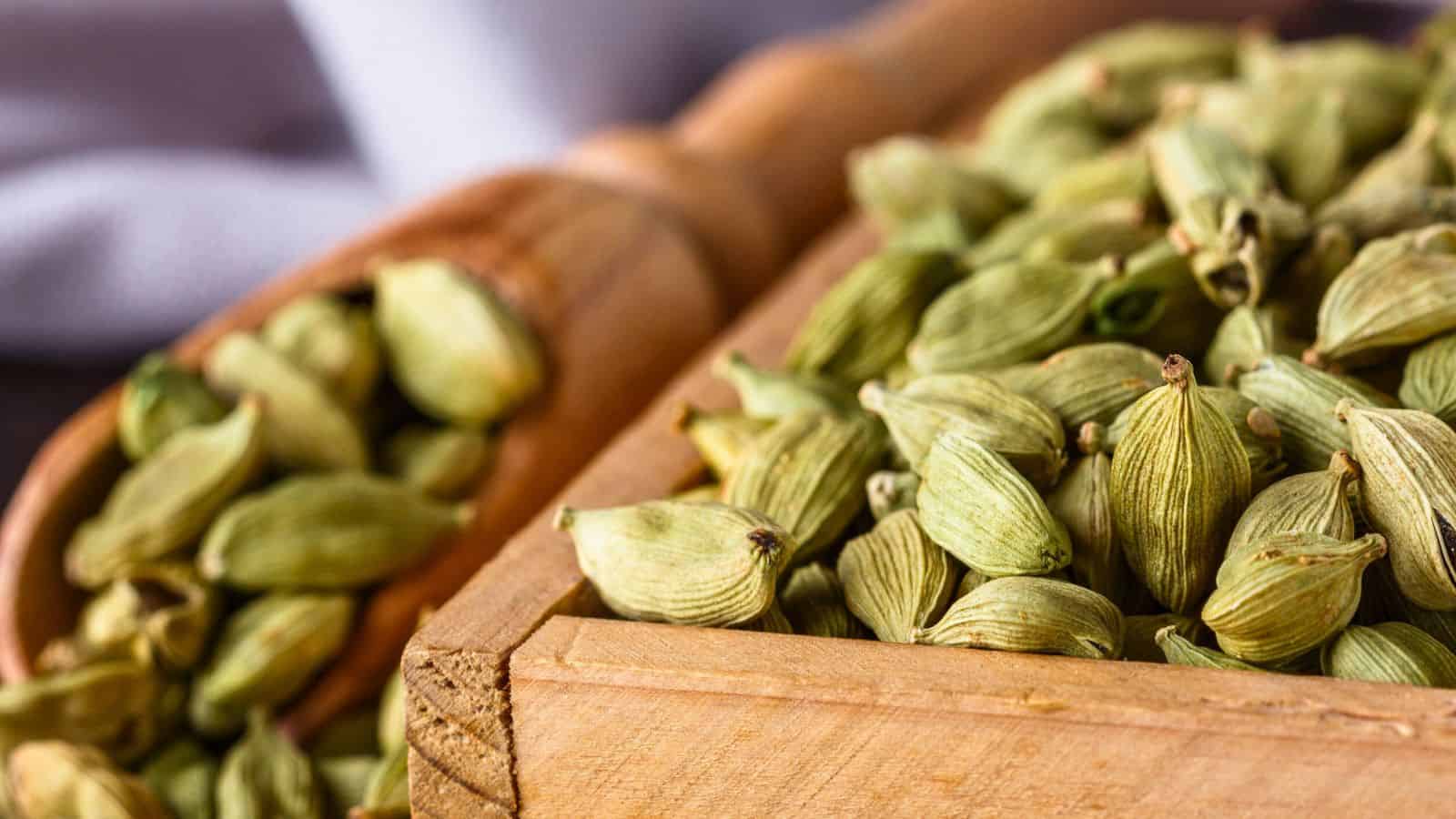 Close-up of a wooden container filled with green cardamom pods. A wooden scoop in the background also holds cardamom pods. The image highlights the texture and natural color of the spice.
