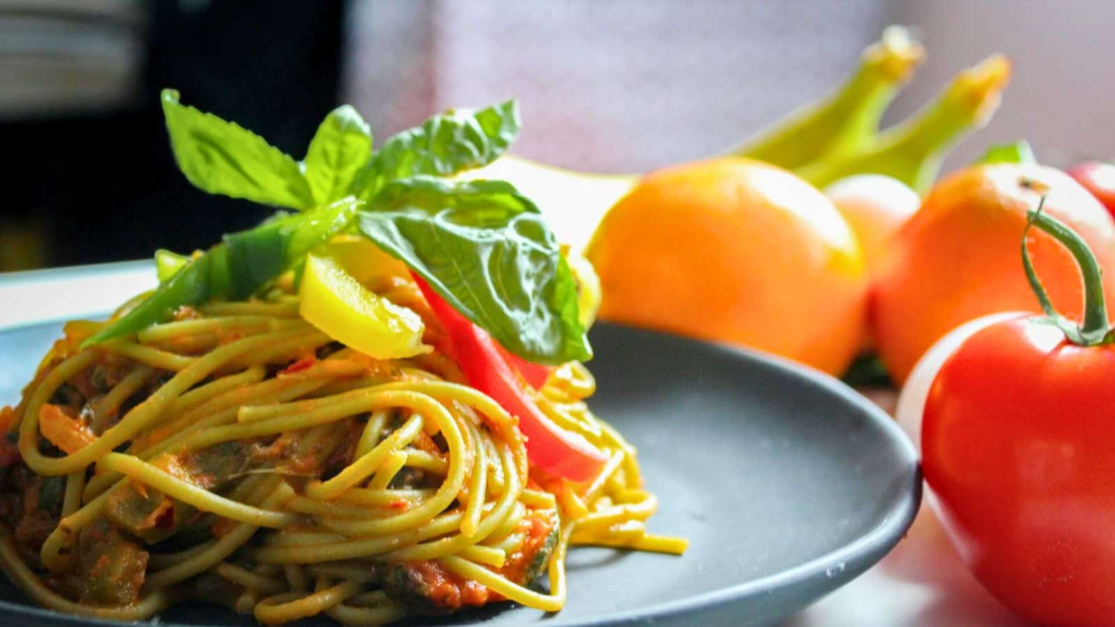 A black plate with spaghetti garnished with basil and bell peppers. In the background, there are oranges and tomatoes, slightly out of focus. The setting appears to be a kitchen or dining area.