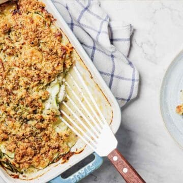 A baked dish of zucchini gratin with a golden breadcrumb topping in a rectangular baking dish. A serving is placed on a round plate next to the dish. A checkered cloth and herbs are in the background on a marble surface.