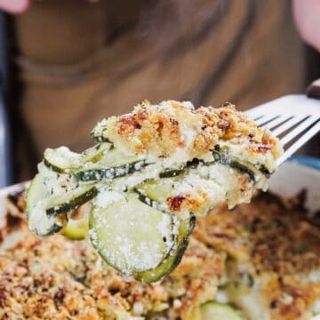 A person holds a serving of zucchini gratin on a spatula above a casserole dish. The gratin has a golden-brown breadcrumb crust and layers of sliced zucchini visible. Steam is rising from the dish, indicating it is freshly baked.