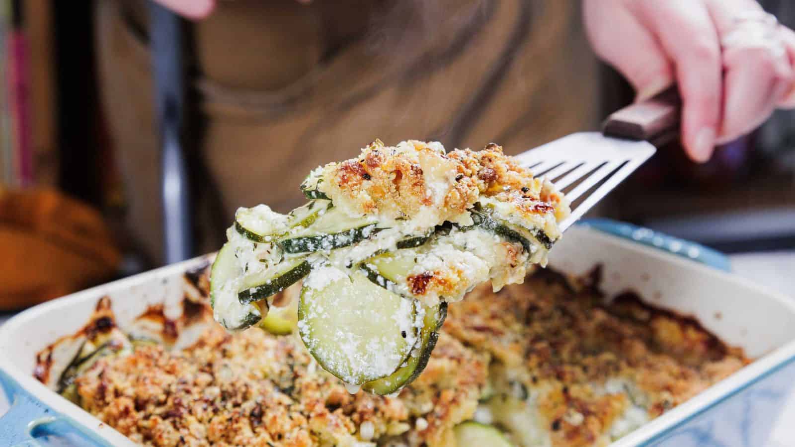 A person holds a serving of zucchini gratin on a spatula above a casserole dish. The gratin has a golden-brown breadcrumb crust and layers of sliced zucchini visible. Steam is rising from the dish, indicating it is freshly baked.