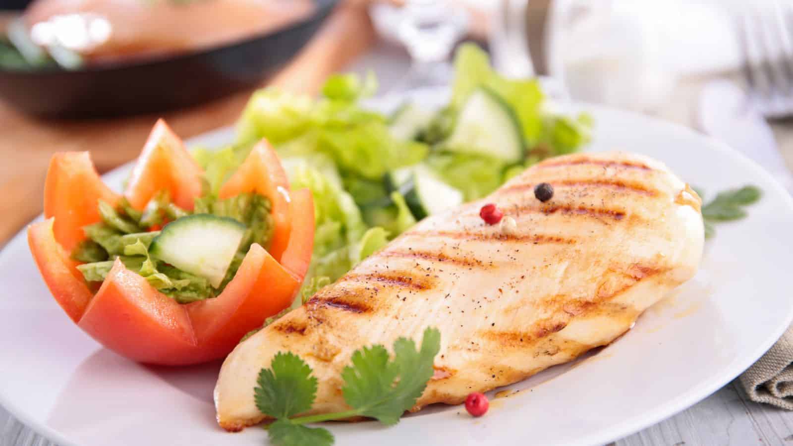 Grilled chicken breast on a white plate, garnished with peppercorns and cilantro, accompanied by a tomato flower filled with lettuce and cucumber slices, and a side of leafy green salad.