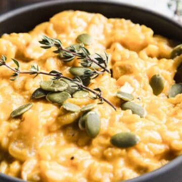 A black bowl filled with creamy, yellow pumpkin risotto. The dish is garnished with fresh thyme sprigs and scattered pumpkin seeds. A spoon rests inside the bowl, and a blurred background shows additional thyme sprigs.
