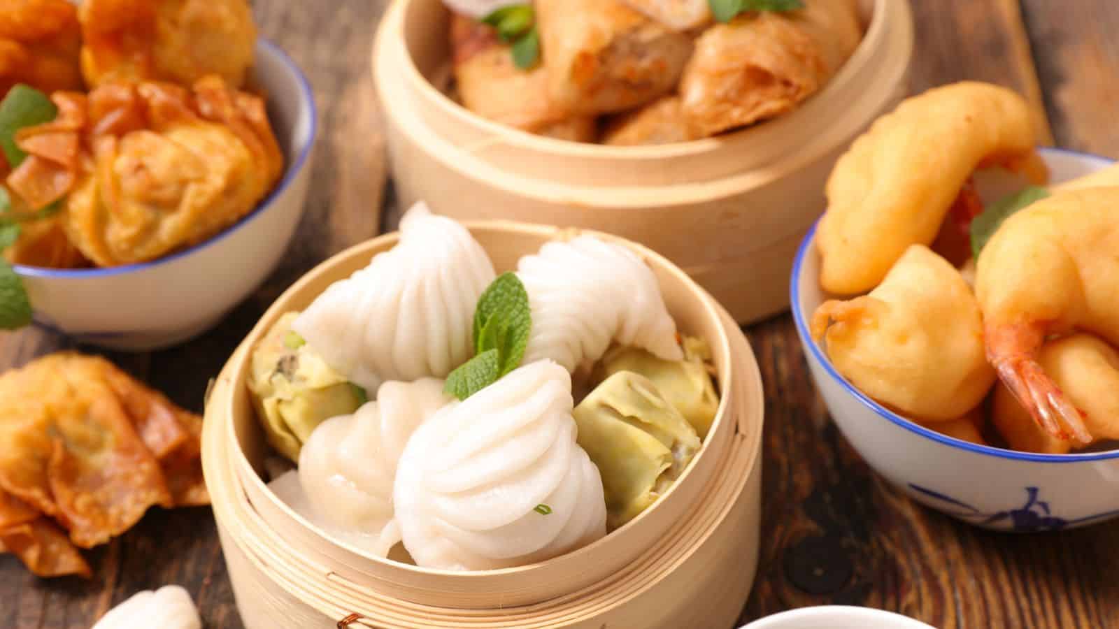 A wooden table displays various Chinese appetizers, including steamed dumplings in a bamboo steamer, crispy fried wontons in a bowl, golden spring rolls, and tempura shrimp in another bowl, garnished with herbs.