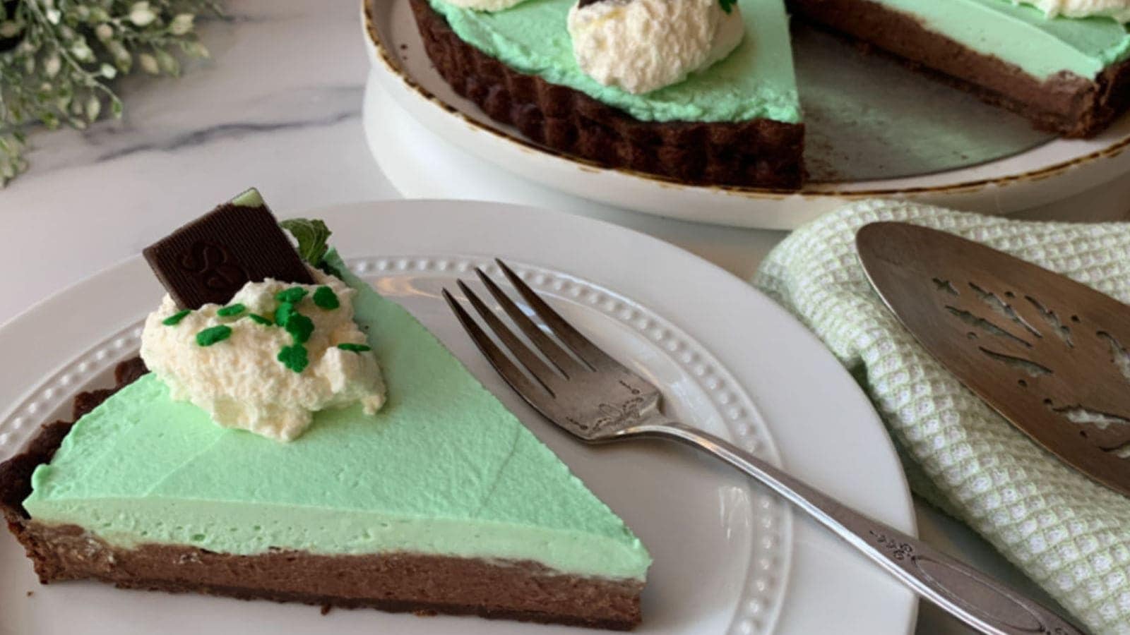 A slice of mint chocolate tart on a white plate with a fork. The tart has a chocolate crust, mint green filling, and is topped with whipped cream, chocolate pieces, and green sprinkles. A serving tool rests nearby on a green cloth.