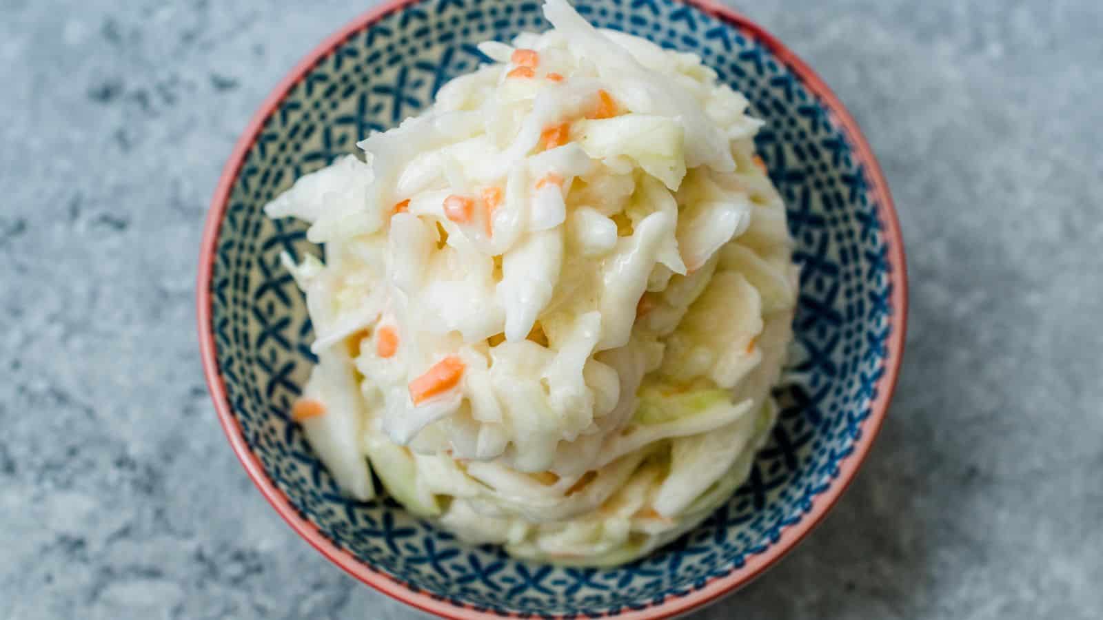A patterned bowl filled with creamy coleslaw, featuring shredded cabbage and small pieces of carrot, sits on a gray surface.