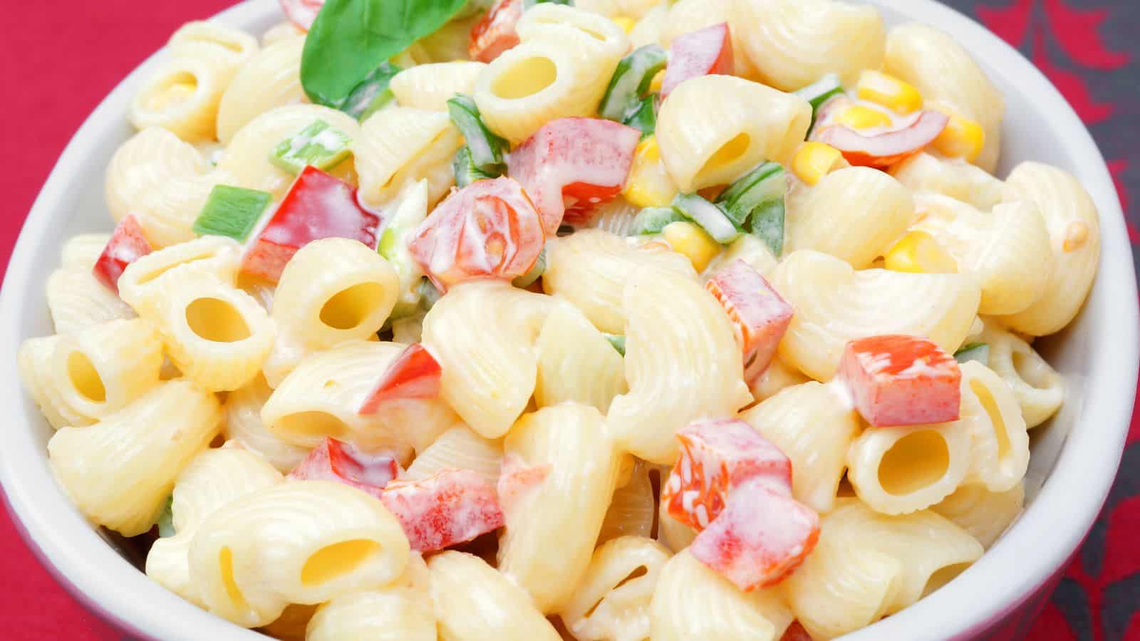 A bowl of macaroni salad featuring elbow pasta mixed with diced red bell pepper, corn, green onions, and a creamy dressing. A basil leaf is used as garnish on top. The bowl is set against a red and black patterned background.