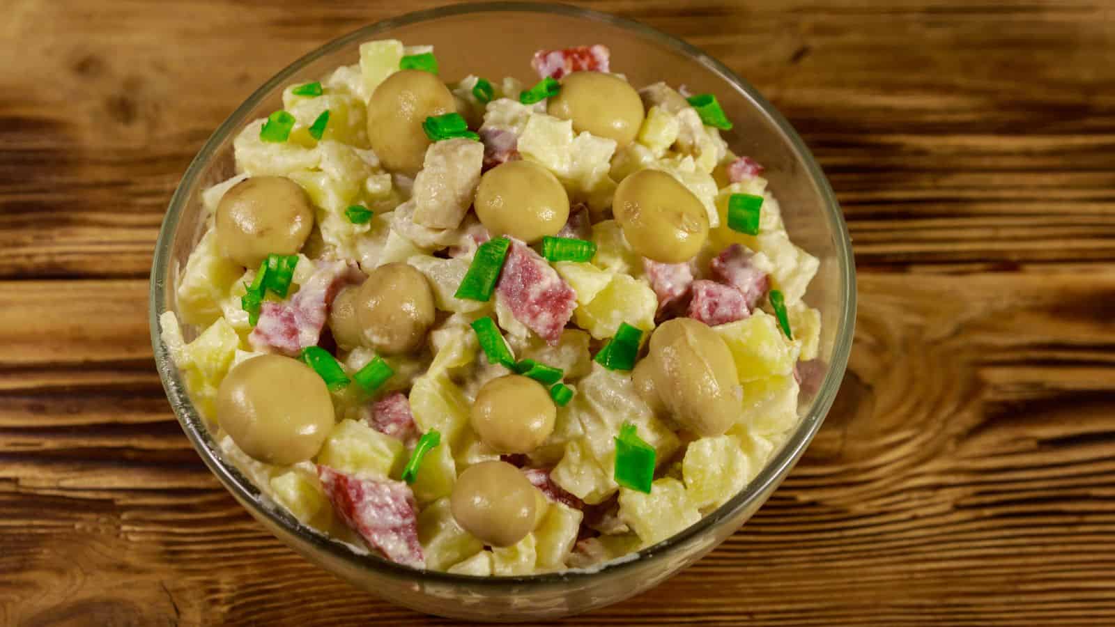 A clear bowl filled with a salad containing diced potatoes, meat, and small brown mushrooms, topped with chopped green onions. The bowl is placed on a wooden surface.