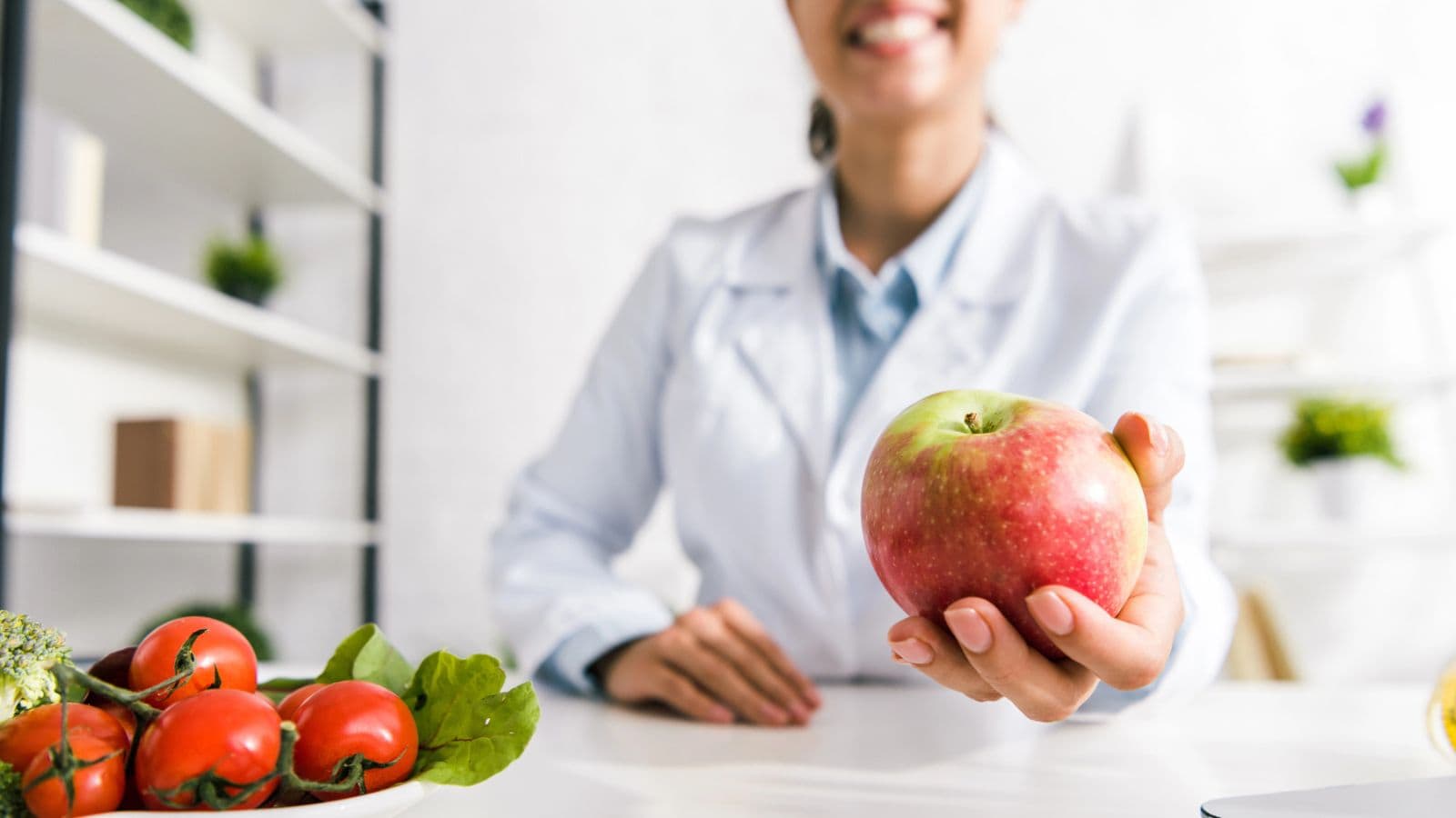 A person in a white coat is holding out a red apple towards the camera. A bowl of assorted vegetables, including tomatoes and lettuce, is on the table. Shelves are visible in the background.