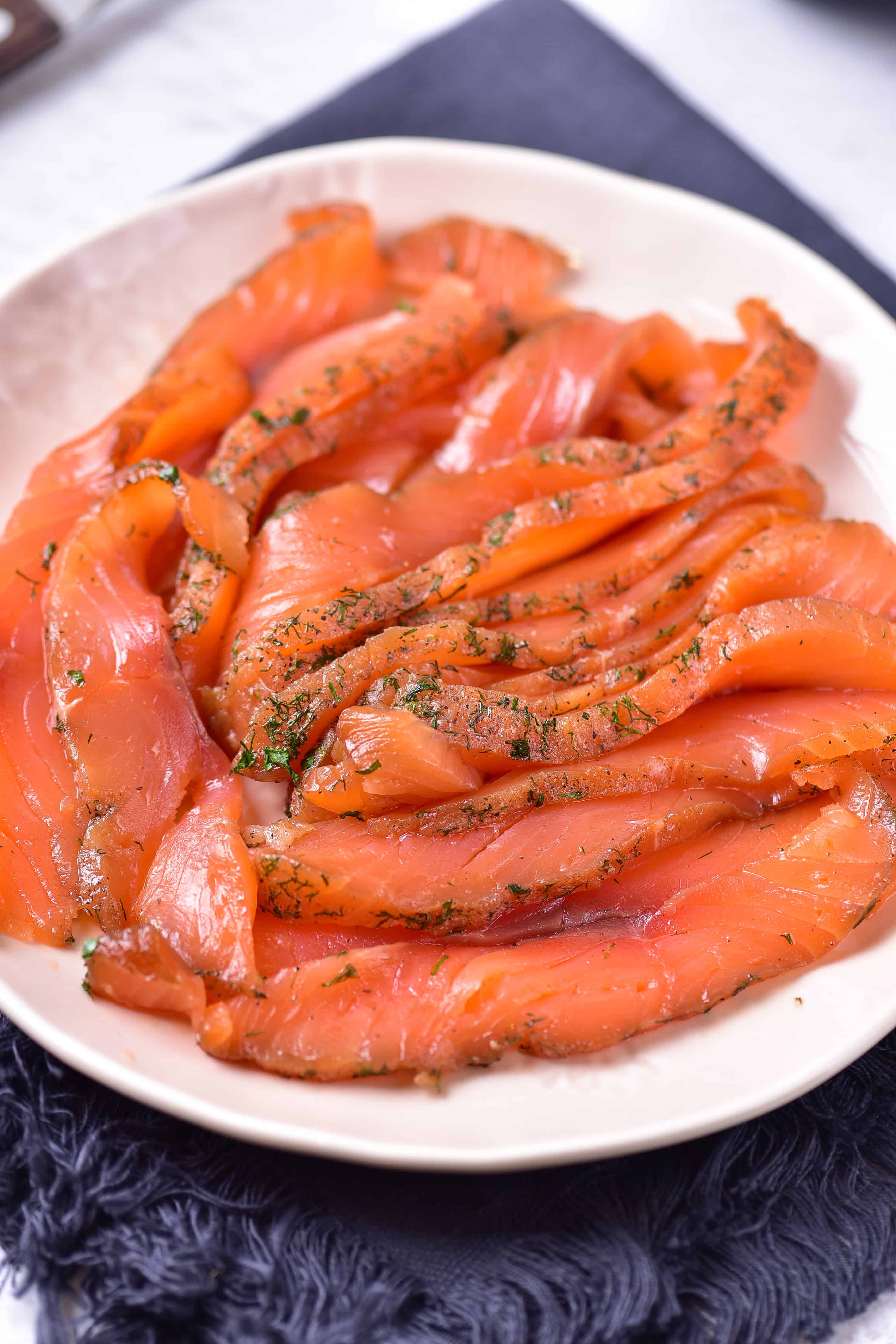 A white plate with several slices of gravlax salmon, garnished with fresh dill, is placed on a dark fabric napkin. The salmon is thinly sliced and boasts a vibrant orange color, making this an easy recipe for an elegant presentation.
