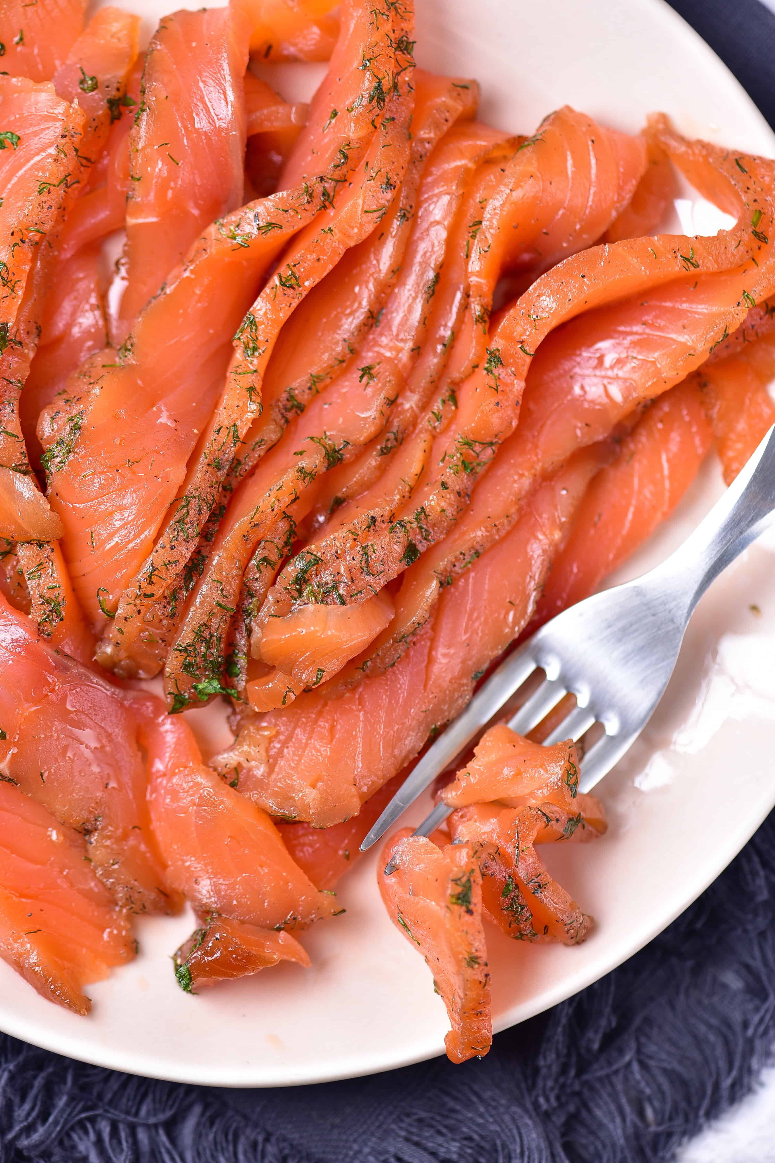 Close-up of a plate with slices of smoked gravlax salmon seasoned with dill and herbs. A fork rests on the plate, holding a small piece of salmon. The plate is set on a dark fabric surface, offering an easy recipe for gourmet delight.