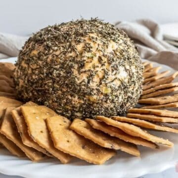 A large, round cheese ball coated in herbs takes center stage on a white plate, promising a crowd-pleasing bite. It's surrounded by neatly arranged crackers, with a folded beige cloth and stacked plates in the background.