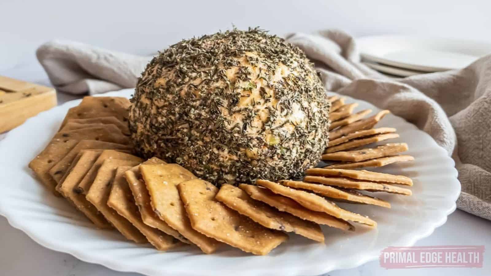 A large, round cheese ball coated in herbs takes center stage on a white plate, promising a crowd-pleasing bite. It's surrounded by neatly arranged crackers, with a folded beige cloth and stacked plates in the background.