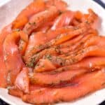 Thin slices of cured salmon, seasoned with dill, are artfully arranged on a white plate. The orange-pink hues of this easy gravlax salmon contrast beautifully with the green flecks of herbs, boasting a texture that appears soft and oily.