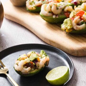 Avocado halves filled with shrimp salad, containing diced tomato, red onion, and herbs, are displayed on a wooden board and black plate. A lime half and a gold fork are placed beside the plate. A wooden bowl with salad is in the background.