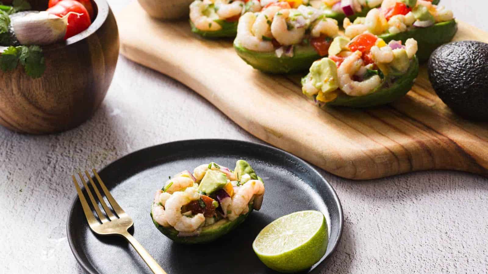 Avocado halves filled with shrimp salad, containing diced tomato, red onion, and herbs, are displayed on a wooden board and black plate. A lime half and a gold fork are placed beside the plate. A wooden bowl with salad is in the background.