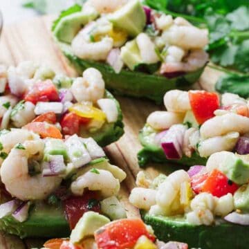 Avocado halves filled with a shrimp salad sit on a wooden surface. The salad includes shrimp, diced tomatoes, red onions, and avocado pieces. A small bowl of salt and a bunch of cilantro are in the background.