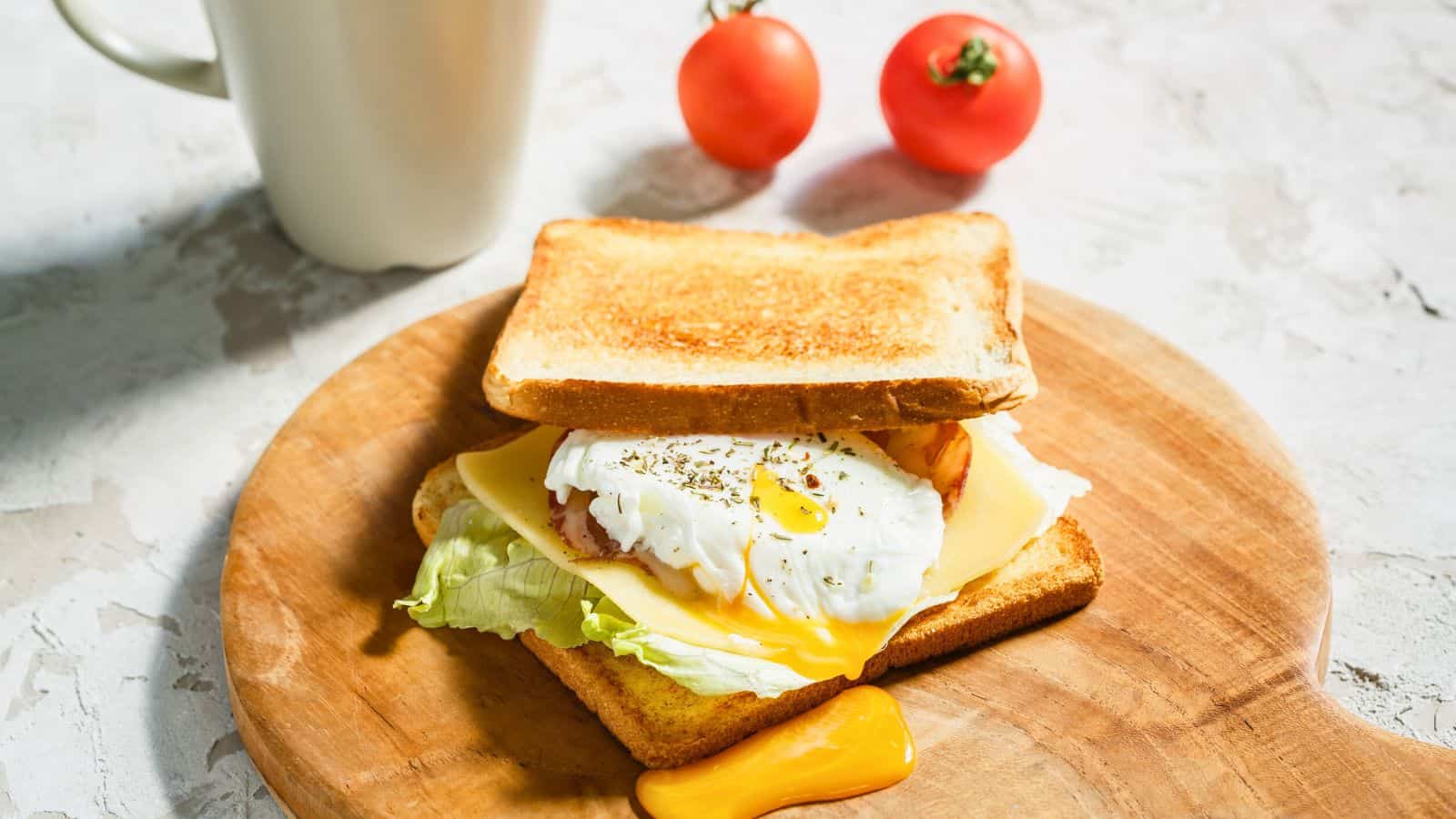 A breakfast sandwich on a wooden board includes lettuce, bacon, cheese, and a poached egg between two slices of toasted bread. A cup and two cherry tomatoes are in the background on a light-colored surface.