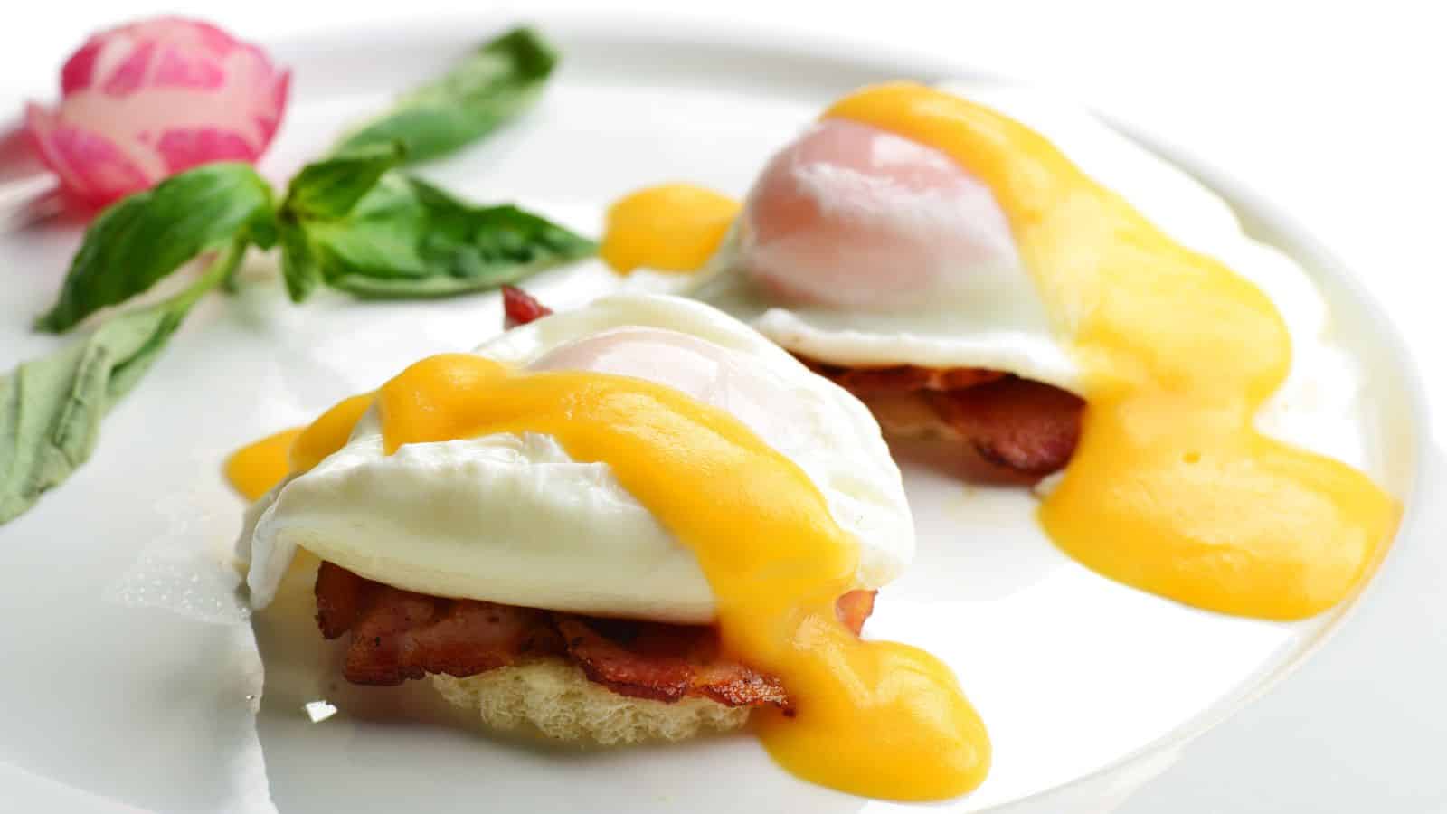 A plate with two eggs Benedict topped with rich yellow hollandaise sauce, resting on slices of bacon and bread. A decorative pink rose and green leaves are placed in the background on the plate.