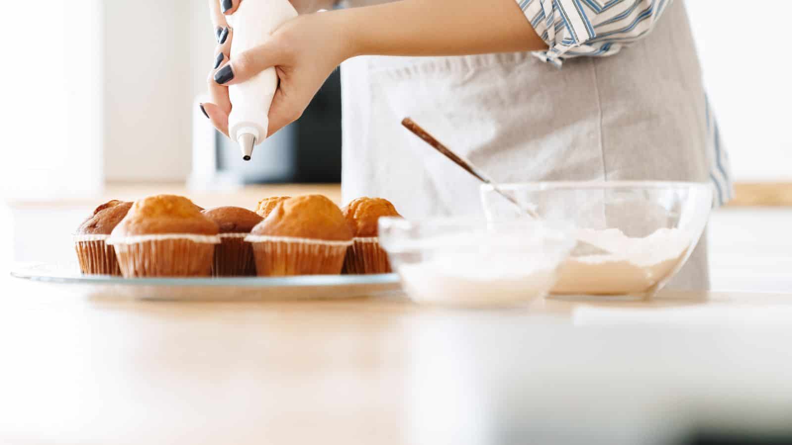 A person wearing an apron is decorating muffins with icing from a piping bag. The muffins are on a plate next to a bowl of flour and a mixing bowl with a spoon. The scene is set in a bright kitchen.