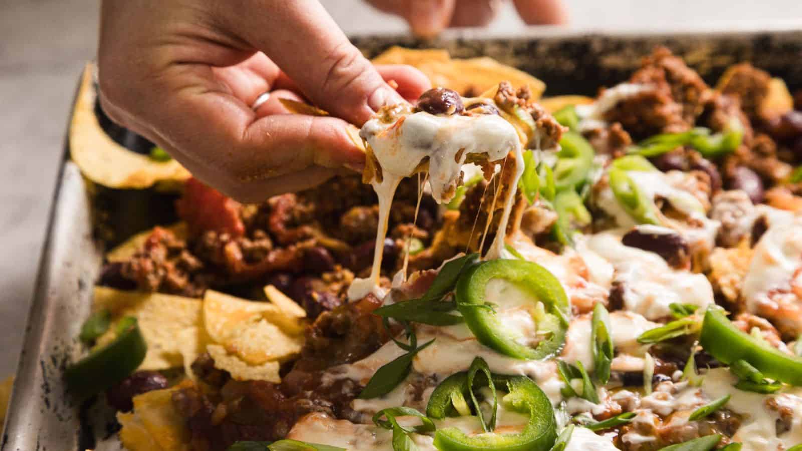 A close-up of nachos topped with melted cheese, ground beef, sliced jalapeños, chopped green onions, sour cream, and beans. The tortilla chips are spread out with the toppings generously applied, creating a colorful and appetizing presentation.