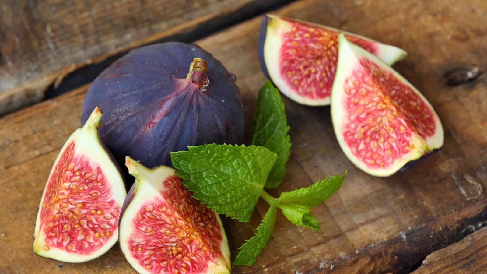 Whole and sliced figs with a dark purple skin and red interior are displayed on a wooden surface alongside a cluster of fresh green mint leaves.