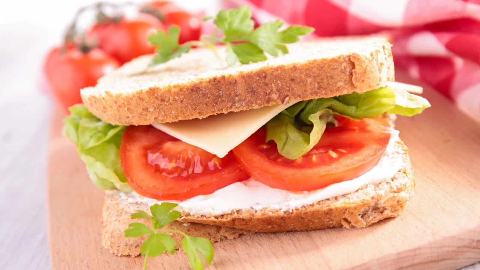 A sandwich with whole-grain bread, containing lettuce, tomato slices, a slice of cheese, and a spread. It is garnished with a parsley leaf. In the background, there are whole tomatoes and a red and white checkered cloth.