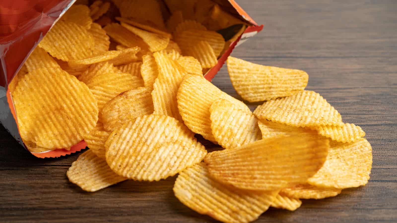 A bag of ridged potato chips is tipped over on a wooden surface, with several chips spilling out onto the table. The chips are golden brown and appear crispy.