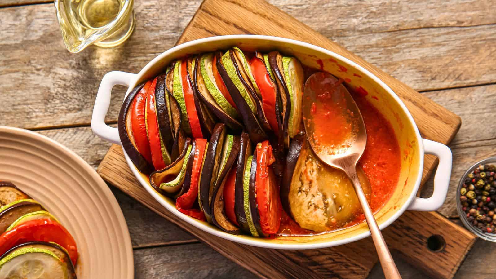 A casserole dish filled with sliced vegetables like zucchini, eggplant, and tomato, arranged vertically. A spoon rests in a sauce on one side. The dish is placed on a wooden surface, with a bowl of peppercorns and a glass jug nearby.